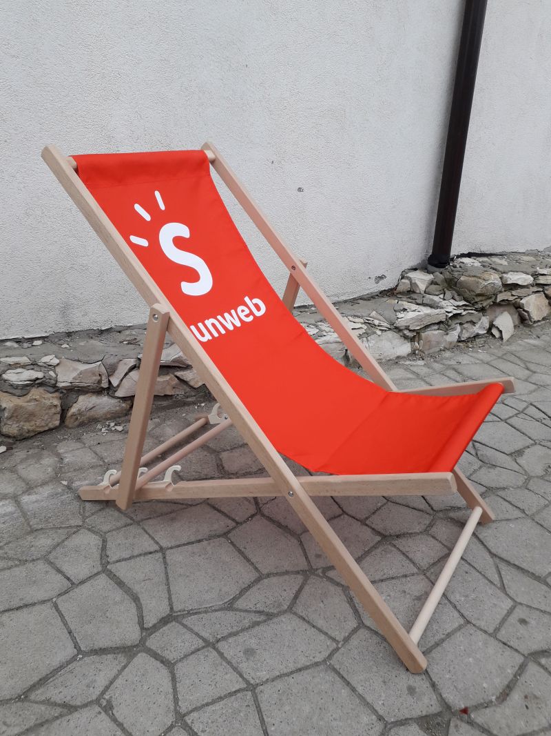 Drewniany leżak plażowy z intensywnie czerwonym siedziskiem i białym logo marki Sunweb, ustawiony na kamiennym podłożu.