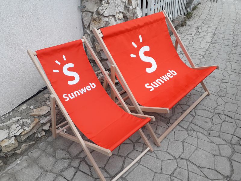 Dwa drewniane leżaki plażowe z czerwonymi siedziskami i białym logo Sunweb, ustawione obok siebie na kamiennym bruku.