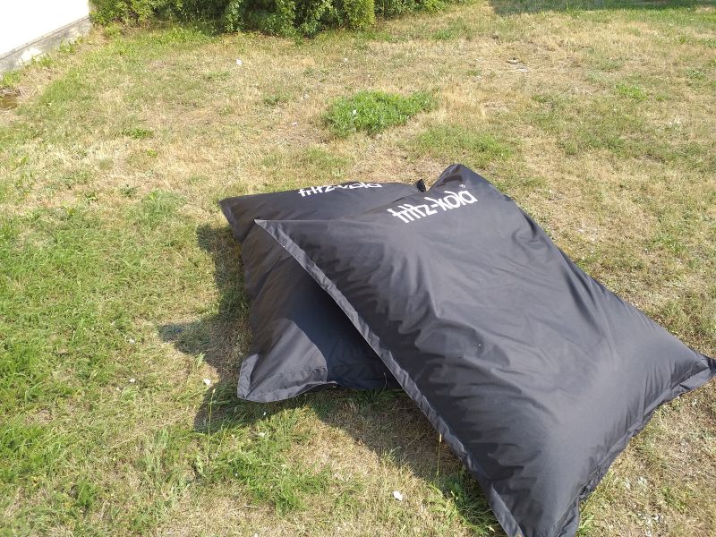 Dwie czarne pufy reklamowe typu „bean bag” z białym logotypem fritz-kola leżące na trawniku.