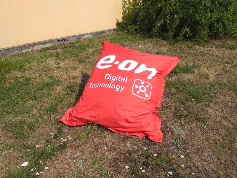 Czerwona pufa reklamowa typu „bean bag” z białym logotypem E.ON Digital Technology leżąca na trawniku.