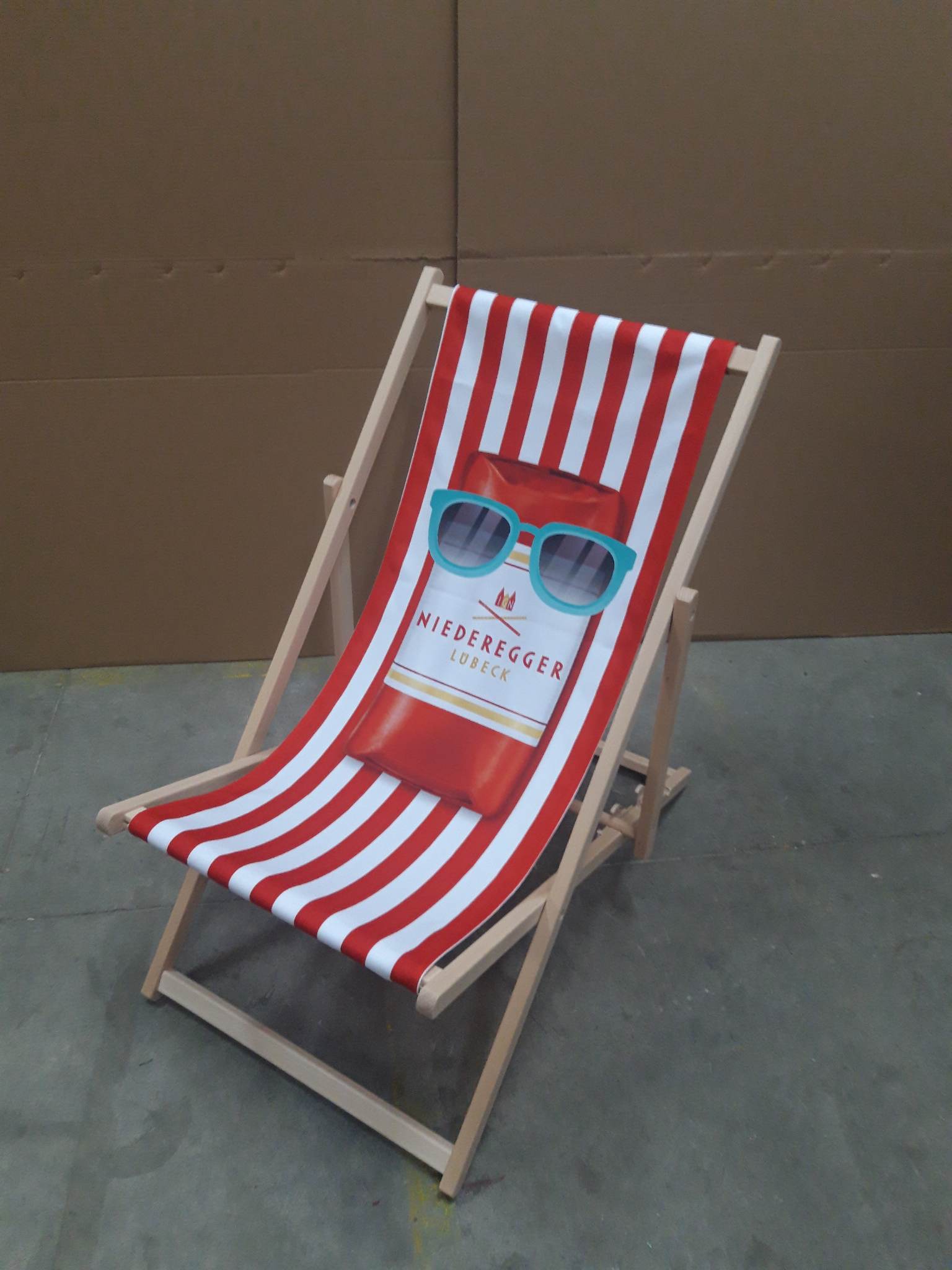Drewniany leżak plażowy z nadrukiem w czerwono-białe paski, ozdobiony grafiką produktu Niederegger Lubeck z okularami przeciwsłonecznymi.