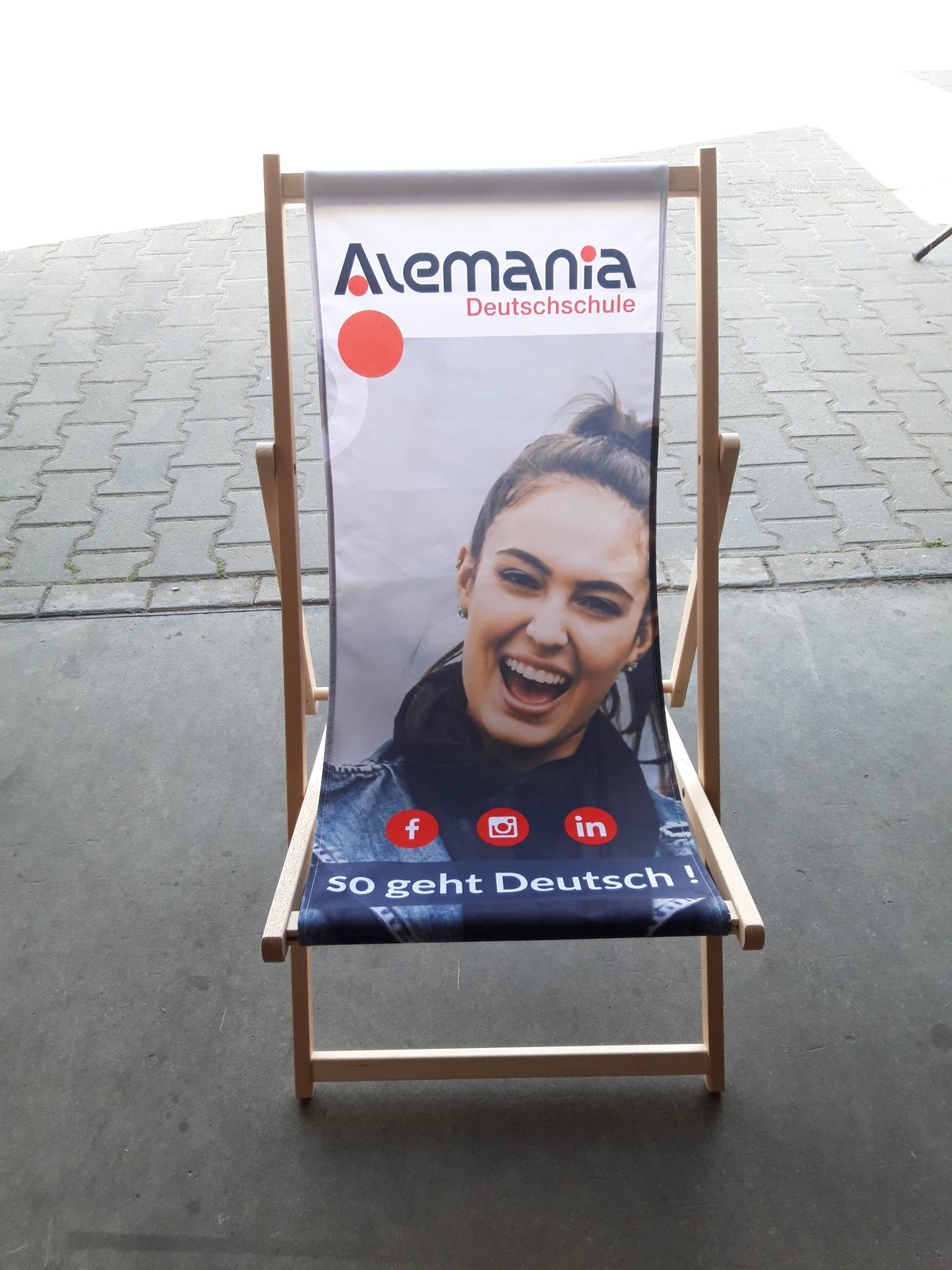 Drewniany leżak plażowy z grafiką szkoły języka niemieckiego "Alemania", przedstawiający uśmiechniętą osobę oraz ikony mediów społecznościowych.