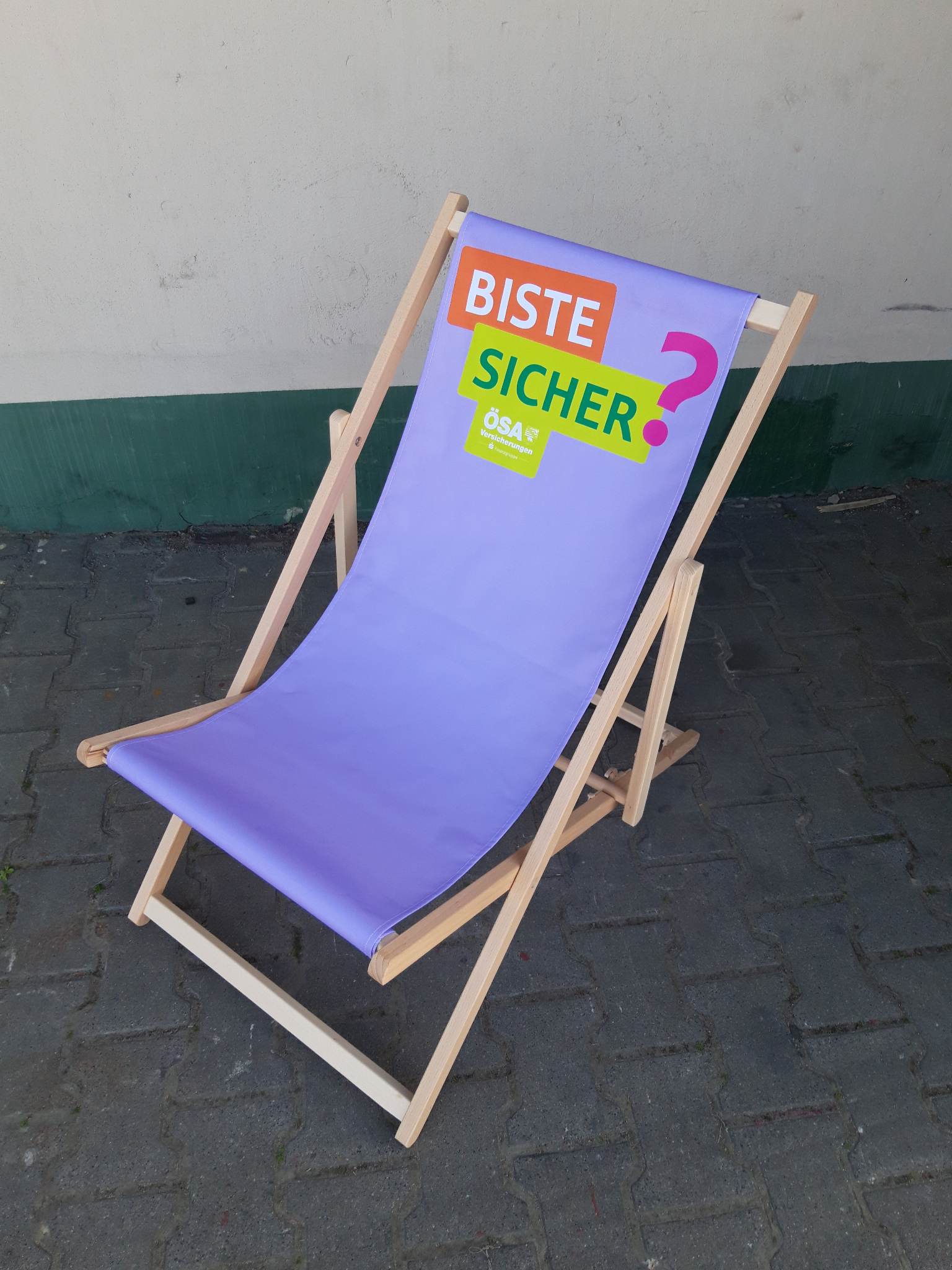 Drewniany leżak plażowy z fioletowym siedziskiem, na którym widnieje napis „BISTE SICHER?” oraz logo ubezpieczalni ÖSA.