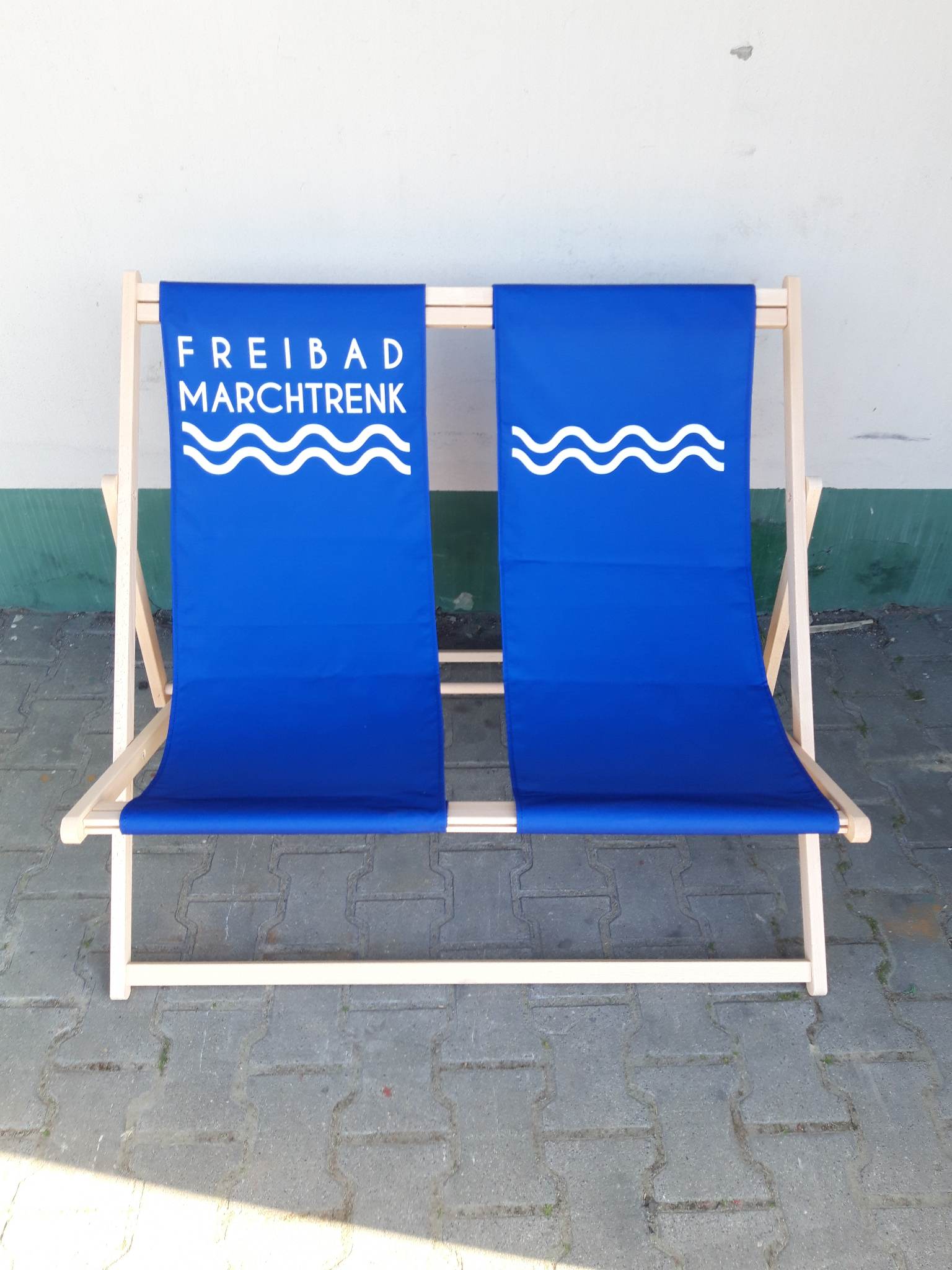 Podwójny leżak plażowy z niebieskim siedziskiem i białym nadrukiem „FREIBAD MARCHTRENK” oraz motywem fal.