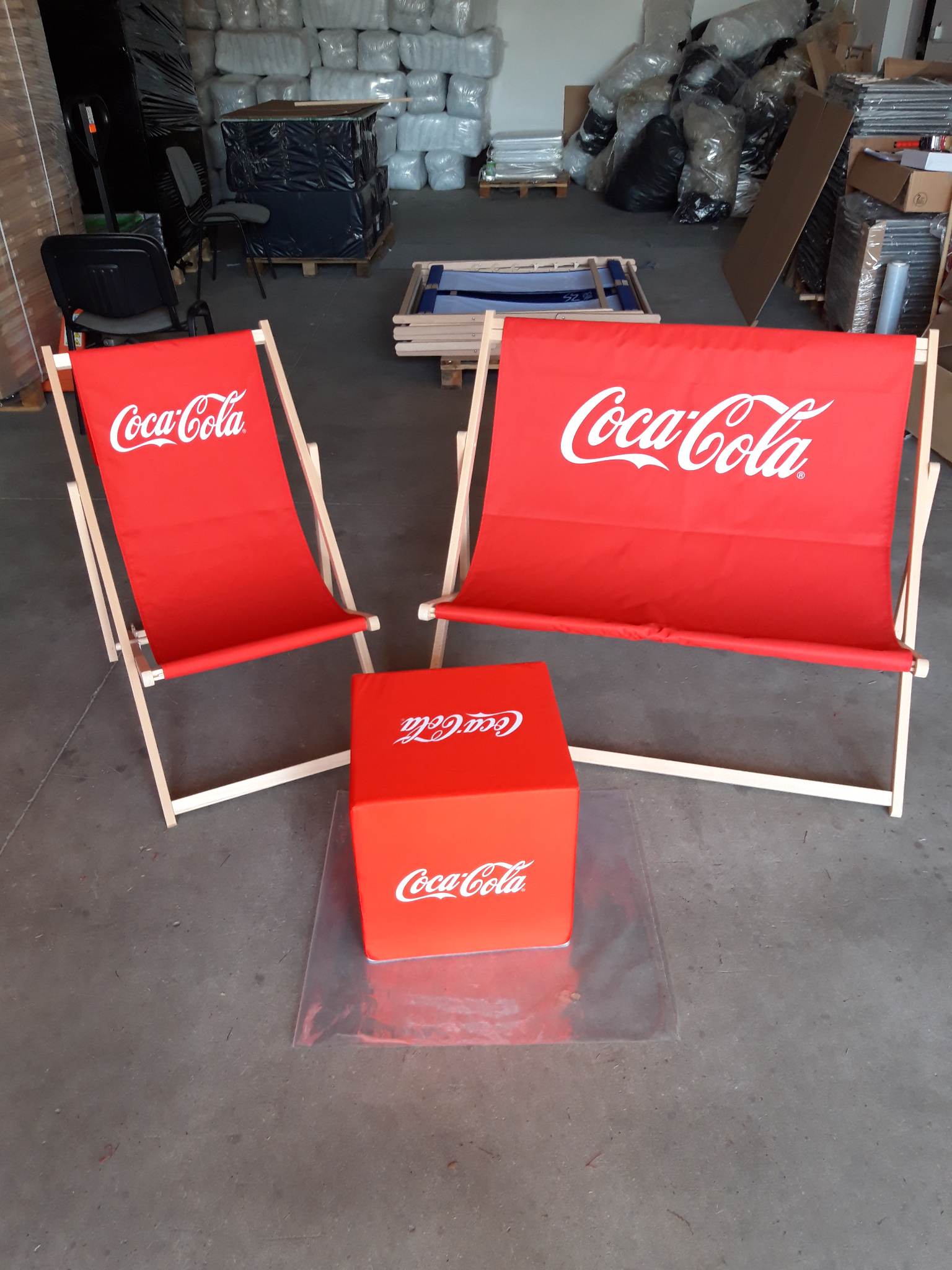 Reklamowy zestaw Coca-Cola obejmujący dwa drewniane leżaki plażowe oraz kwadratową pufę, wszystko w charakterystycznym czerwonym kolorze z białym logo.