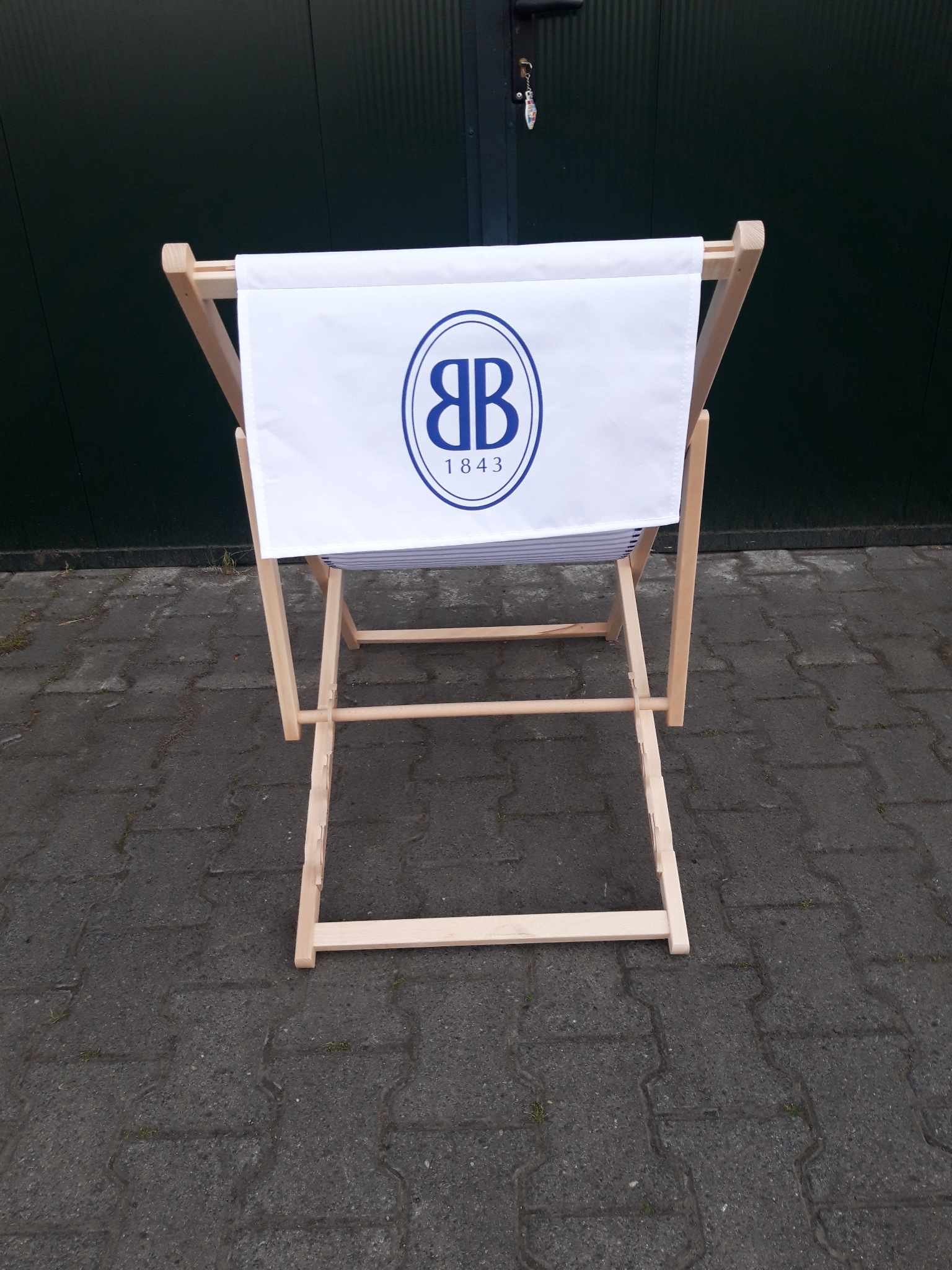 Drewniany leżak plażowy widziany od tyłu, z białą tkaniną ozdobioną niebieskim logo „BB 1843” w owalnej ramce.