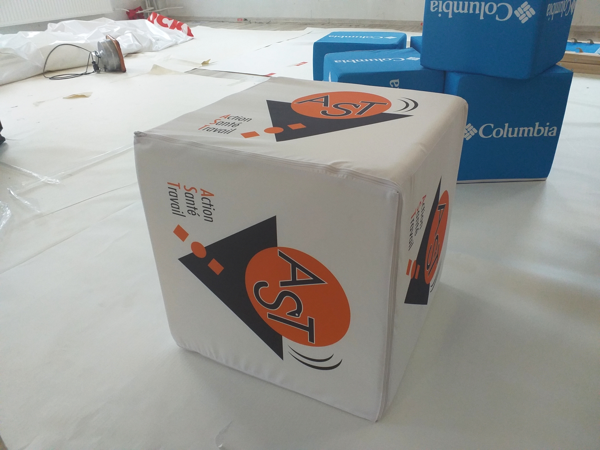 Reklamowa pufa w kształcie kostki o wymiarach 50x50x50 cm z logo AST (Action Santé Travail) oraz tło z niebieskimi kostkami marki Columbia.