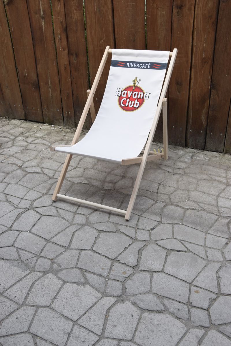 Drewniany leżak plażowy z białym siedziskiem, na którym widnieje logo marki Havana Club oraz napis RIVERCAFÉ, ustawiony na kamiennym bruku przy drewnianym płocie.