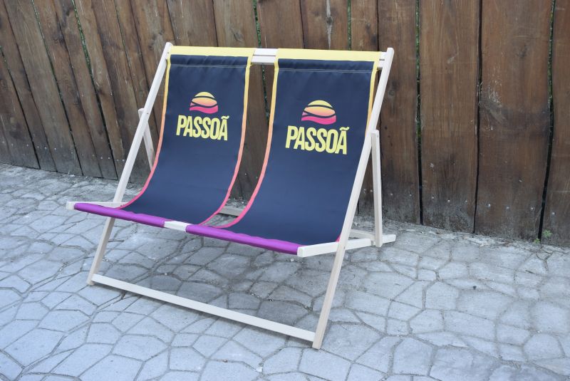 Podwójny leżak plażowy z drewnianą konstrukcją i czarną tkaniną z nadrukiem logo Passoa, ustawiony na kamiennym bruku.