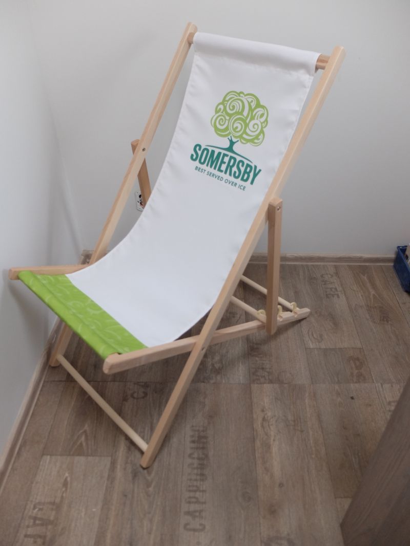 Drewniany leżak plażowy z białym siedziskiem z zielonym logo marki Somersby, ustawiony w pomieszczeniu na drewnianej podłodze.