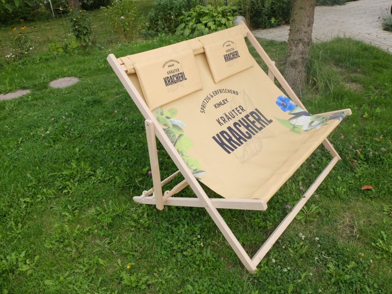 Podwójny leżak plażowy z beżową tkaniną, grafiką ziołową i logotypem Kinley Kräuter Kracherl, ustawiony na trawniku.