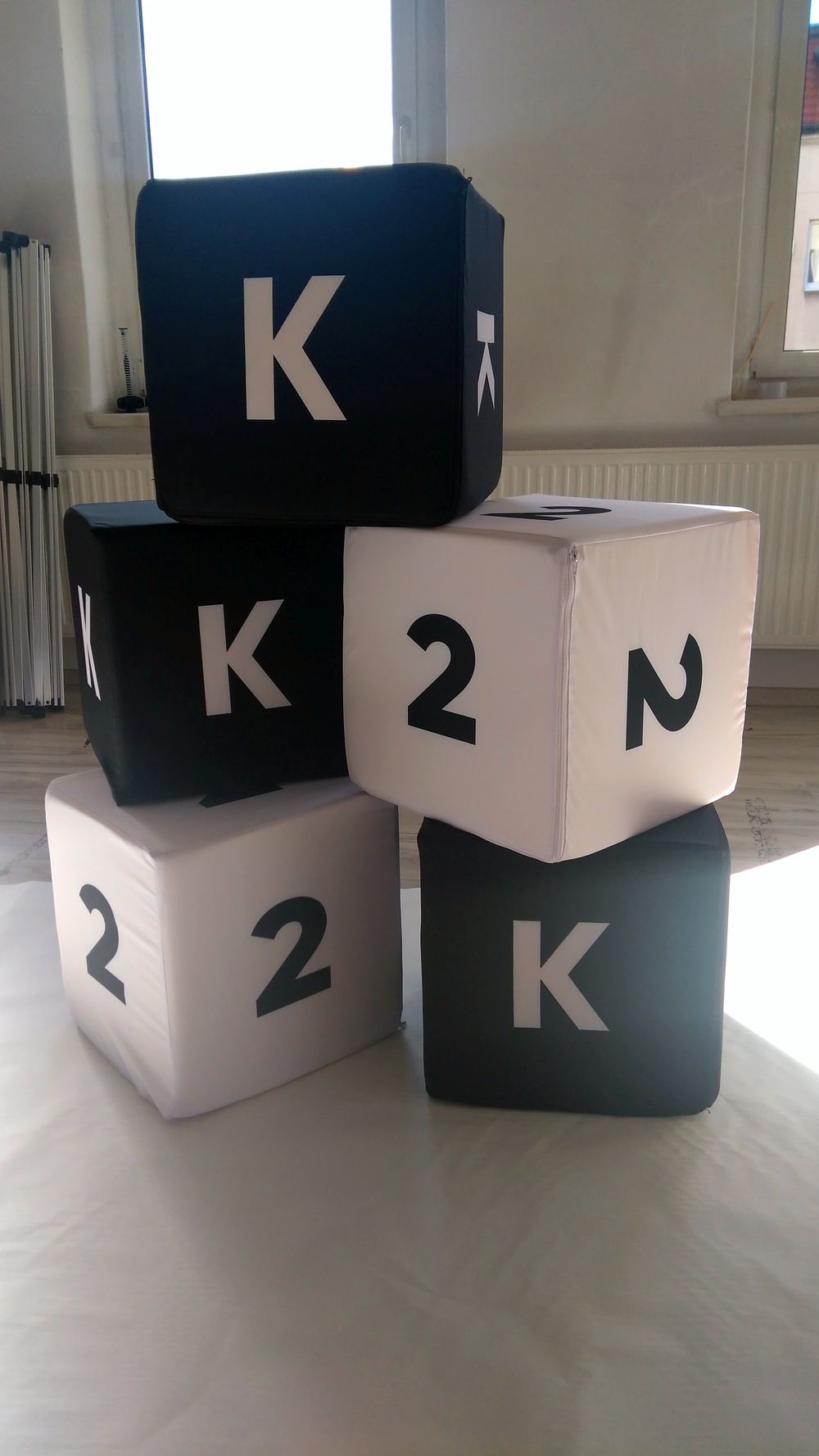 Zestaw czarno-białych puf reklamowych w kształcie kostek z nadrukowanymi literami „K” oraz cyframi „2”.