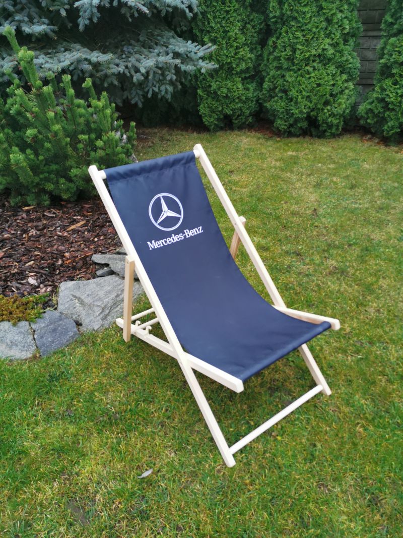 Drewniany leżak plażowy z ciemnogranatowym siedziskiem i białym logo Mercedes-Benz, ustawiony na tle zielonego ogrodu.