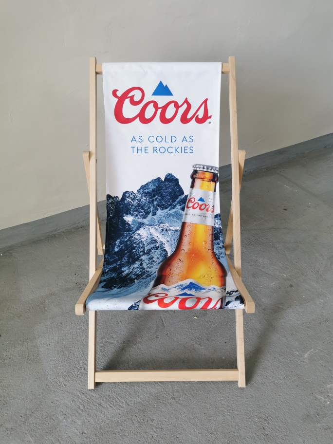 Drewniany leżak plażowy z nadrukiem marki piwa Coors, grafiką ośnieżonych gór oraz butelką na białym tle.