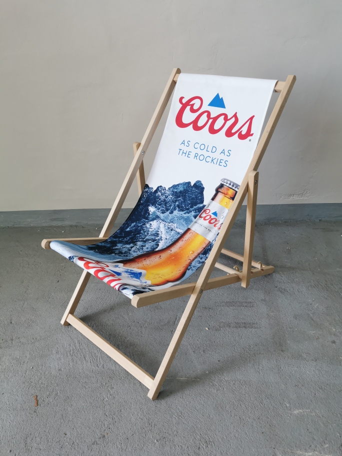 Drewniany leżak plażowy z widocznym brandingiem piwa Coors, grafiką ośnieżonych gór oraz butelką na białym tle, ustawiony wewnątrz budynku.