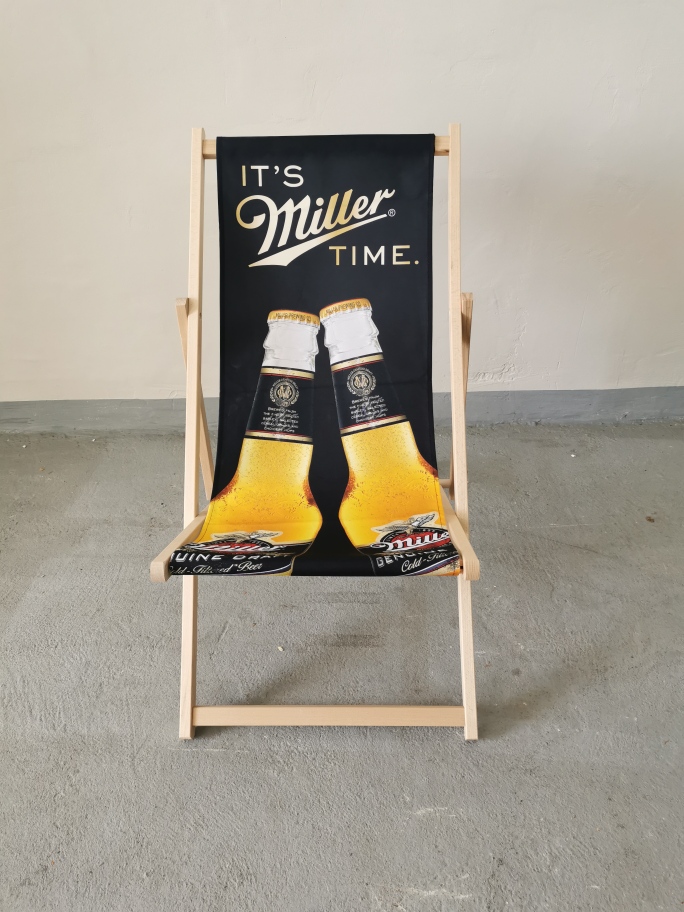 Drewniany leżak plażowy z czarnym siedziskiem, na którym widnieje logo "It's Miller Time" oraz grafika dwóch butelek piwa.