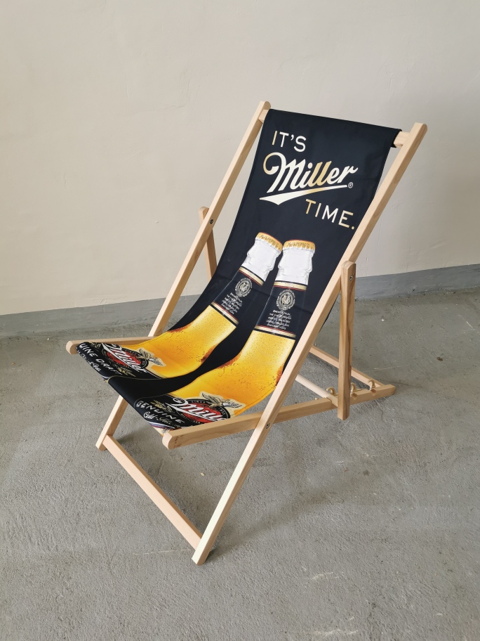 Drewniany leżak plażowy z czarnym siedziskiem, hasłem "It's Miller Time" oraz grafiką dwóch butelek piwa, ustawiony wewnątrz budynku.